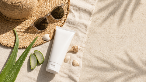 Tubo de crema solar sin marca, sombrero de paja, gafas de sol y hoja de aloe vera sobre fondo claro de arena y lino.