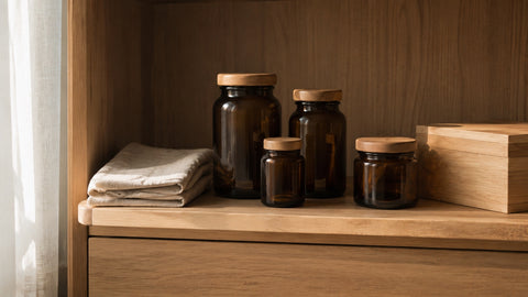 Balda de un armario de madera clara con cuatro frascos de cristal ámbar sin etiqueta y tapón de madera, junto a un paño de lino doblado y una caja de madera