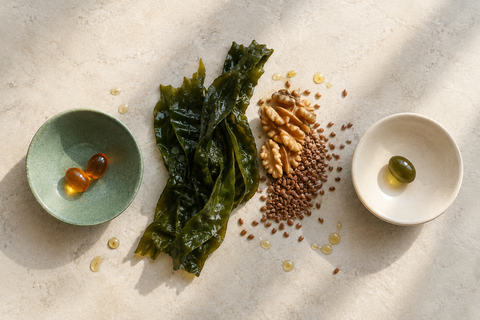 Dos cuencos de cerámica, uno con perlas ámbar de aceite de krill y otro con una perla verde de omega 3 vegano, junto a alga fresca, nueces y semillas de lino.